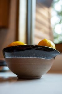 a clay coyote cassole bowl in natural clay and midnight black rim is viewed from the side, the viewpoint is from the kitchen countertop with the rest of the visible room out of focus in the background. There are two lemons just peaking out of the top of the bowl.
