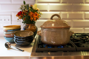 a horizontally framed picture. a clay coyote dutch oven is sitting on the iron grate of a stove top gas burner range. The burner is lit and there is a small blue flame visible underneath the dutch oven. The dutch oven is in coyote grey with darker speckles through out the glaze. The lid is on, and the handles are pointing to the left and right sides of the frame. To the left of the dutch ovens are two stacks of clay coyote chili bowls. the nearest stack of bowls is 3 high with dark glazes visible, and the top bowl is in mint chip glaze. The second stack of bowls, slightly behind the first and to the left is 5 high with 2 joes blue, 1 mint chip and a yellow salt glaze on the top. the bottom bowl is hard to tell glaze. Behind the stack of first bowls is a clay coyote large vase in joes blue, filled with a fall bouquet with orange, yellow and red flowers