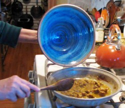 phot of a person taking a blue tagine lid off of the tagine and the stove and stirring whats in it