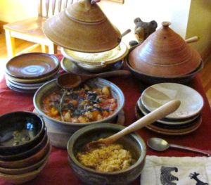 photo of multiple tagines on a table with a stack of plates and bowls and a bowl of couscous with a wooden spoon in it