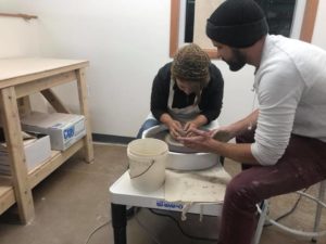 photo of a woman sculpting a pot on the pottery wheel with a man helping her