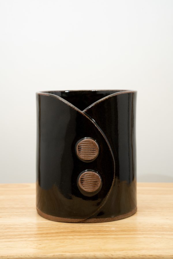 the photograph shows a clay coyote utensil holder and wine chiller glazed in midnight black. it is sitting on a wooden surface. the wooden surface has a light wood color to it. the utensil holder and wine chiller has two decorative buttons on the front of it. they are unglazed and show off the natural reddish brown of the clay. each button has a horizontal line motif on them. the background is a plain white wall. the photograph is well lit with white light.