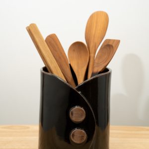 the pictures shows a clay coyote utensil holder and wine chiller glazed in black. it has 4 Jonathan's spoons utensils and one Baer wooden tongs resting inside of it. the utensils are all available for sale at clay coyote. from left to right it goes: baer wooden tongs, Jonathan's spoons wooden spatula, 2 Jonathan's spoons spoons and finally a Jonathan's spoons combo spoon and spatula. the utensil holder and wine chiller has two decorative buttons on the front of it, each one has horizontal stripes across their faces. the faces are unglazed show the natural reddish brown of the clay.