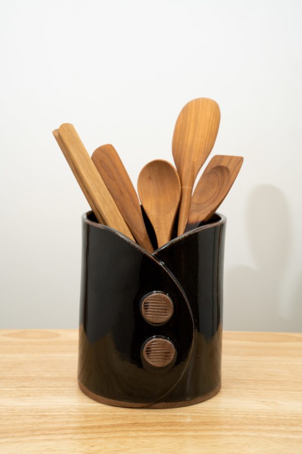 the pictures shows a clay coyote utensil holder and wine chiller glazed in black. it has 4 Jonathan's spoons utensils and one Baer wooden tongs resting inside of it. the utensils are all available for sale at clay coyote. from left to right it goes: baer wooden tongs, Jonathan's spoons wooden spatula, 2 Jonathan's spoons spoons and finally a Jonathan's spoons combo spoon and spatula. the utensil holder and wine chiller has two decorative buttons on the front of it, each one has horizontal stripes across their faces. the faces are unglazed show the natural reddish brown of the clay.
