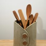 the photograph shows a clay coyote utensil holder and wine chiller glazed in mint. the utensil holder and wine chiller is holding 5 utensils. it is holding from left to right: a Jonathan's spoons spatula/spoon hybrid, a wooden set of baer brand tongs, 2 large Jonathan's spoons spoons and a Jonathan's spoons spatula. all utensils are wooden. the background is a plain white wall. the photograph is well lit with white light.