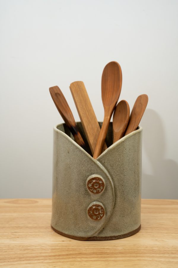 the photograph shows a clay coyote utensil holder and wine chiller glazed in mint. the utensil holder and wine chiller is holding 5 utensils. it is holding from left to right: a Jonathan's spoons spatula/spoon hybrid, a wooden set of baer brand tongs, 2 large Jonathan's spoons spoons and a Jonathan's spoons spatula. all utensils are wooden. the background is a plain white wall. the photograph is well lit with white light.
