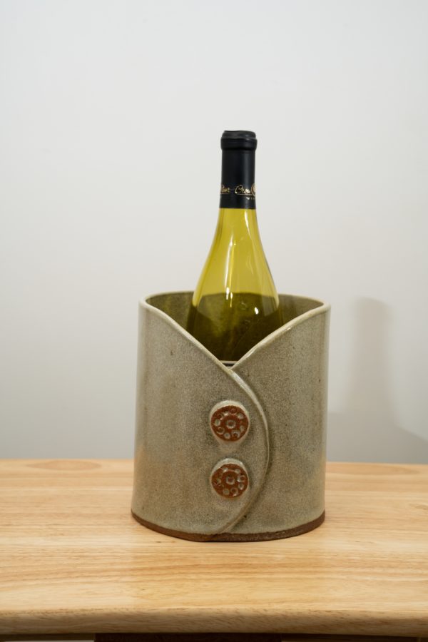 the photograph shows a clay coyote utensil holder and wine chiller glazed in mint. it is sitting on a wooden surface that has light colored wood. the utensil holder and wine chiller has a empty wine bottle in it to show the fit of a standard wine bottle in it. the utensil holder and wine chiller has two decorative buttons on the front of it. the decorative buttons have a circle of small circles, with one small circle in the middle. the background is a plain white wall.