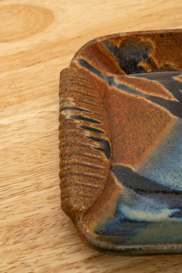 the photograph shows a very close up view of the handle section of a clay coyote small tray glazed in joes blue. the handle is in the brown section of the joes blue glaze. the small tray is resting on a light colored wooden surface. the photograph is lit with white light.