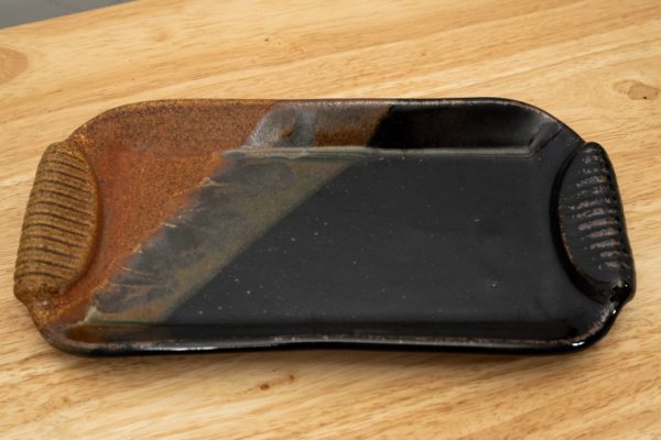 the photograph shows a clay coyote small tray glazed in mocha swirl resting on a light colored wooden surface. the photograph is lit with white light. the brown section of the glaze is pointed towards the left of the photograph.