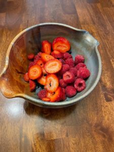a vertically framed photograph shows an overhead view of a clay coyote mixing bowl with strawberries, blueberries and raspberries in it. the strawberries have been sliced. the mixing bowl is glazed in joes blue. the mixing bowl is sitting on a wooden table.