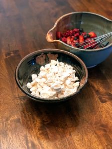 a vertically framed photograph shows a fairly close view of two bowls. One bowl has crushed meringues in it. the bowl is glazed black with little bits of brown. behind and to the right is a clay coyote mixing bowl glazed in joes blue. the mixing bowl has cut up strawberries, blue berries and raspberries in it. there is also a metal wire whisk in the bowl. both of the bowls are resting on a wooden table.