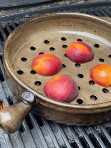 a vertically framed photograph shows a close up high angle view of a clay coyote grill basket grilling 4 peach halves. the peach halves are all cut side down in the grill basket. the peaches still have their skins on. the grill basket has developed a nice patina with usage. the photograph is well lit with natural light.