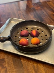 a vertically framed photograph shows a fairly close up view of a clay coyote grill basket that is going to be used to grill 4 peach halves. the peaches are all cut side down on the grill basket, arranged so none of them are touching. the grill basket is resting on a baking sheet, to prevent any drips from the peaches when they are moved from the wooden table they are resting on to the grill.