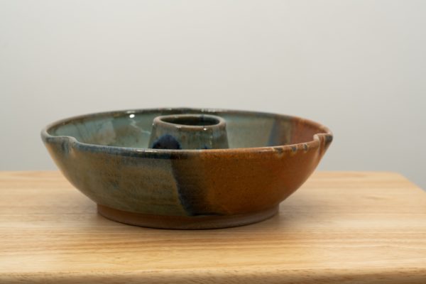 the photograph shows a clay coyote chicken baker glazed in joes blue. the chicken baker is resting on a small light colored wooden table. the photograph is lit with white light. the background is a white wall.