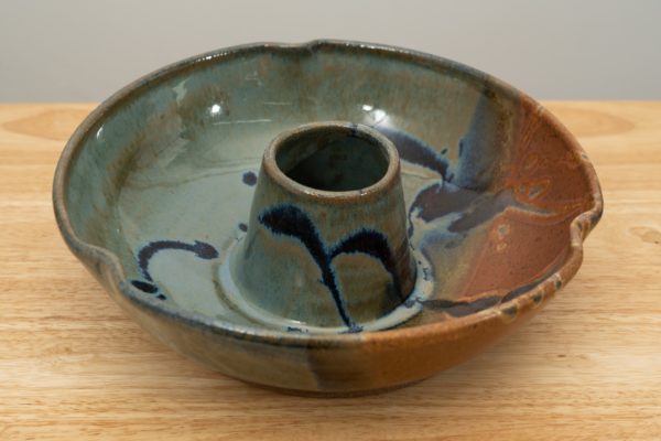 the photograph shows a clay coyote chicken baker glazed in joes blue resting on a light colored wooden surface. the background is a white wall. the photograph is well lit with white light.