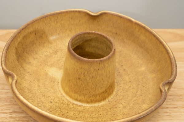 the photograph shows a clay coyote chicken baker glazed in yellow salt resting on a light colored wooden table. the background is a white wall. the chicken baker has been propped up slightly to better show the glaze pattern on the inside of the chicken baker. the photograph is well lit with white light.