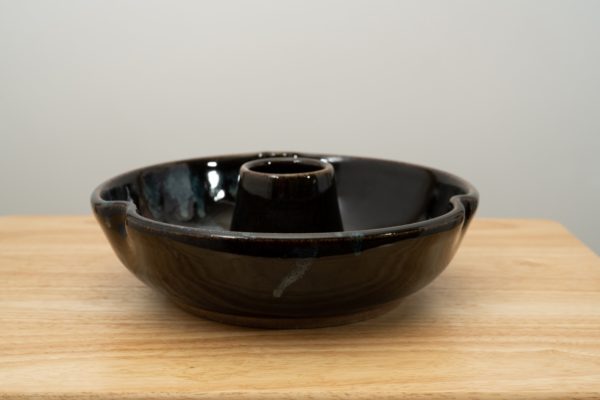 the photograph shows a clay coyote chicken baker glazed in midnight garden resting on a small light colored wooden table. the background is a white wall. the photograph is well lit with white light.