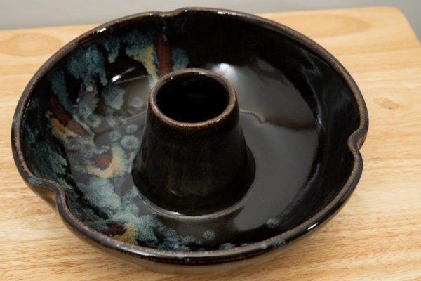 the photograph shows a high angle view of a clay coyote chicken baker glazed in midnight garden resting on a light colored wooden surface. The angle of the the photograph allows the viewer to see inside the chicken baker to see the glaze pattern within. the photograph is well lit with white light.