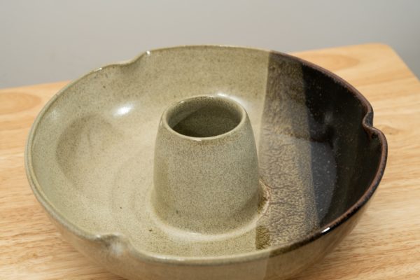 the photograph shows a high angle view of a clay coyote chicken baker glazed in mint chip. the chicken baker is resting on a small light colored wooden table. the angle of the photographs height allows the viewer to see into bowl part of the chicken baker, and to see the glaze pattern on the bottom. the photograph is well lit with white light.