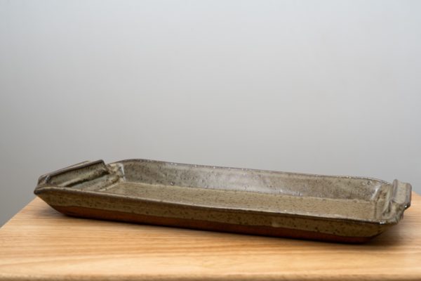 the photograph shows a clay coyote flameware fish tray resting on a small light colored wooden table. the tray is laying flat on the table. the flameware fish tray is glazed in coyote grey (grey with dark speckling through out). the background is a white wall. the photograph is lit with white light.