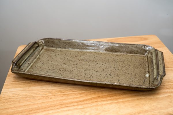 the photograph shows a clay coyote flameware fish tray resting on a small light colored wooden table. the fish tray is glazed in coyote grey (grey with darker speckling through out) the tray is resting flat on the wooden surface. the tray is slightly angled with the left end of the tray pointed towards the left corner of the photograph. the photograph is lit with white light.