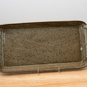 the photograph shows a clay coyote flameware fish tray resting on a stand. the stand allows the fish tray to be almost vertical, and allows the viewer to see the entire inside cooking surface of the tray. the tray and stand are sitting on a small light colored wooden table. the tray is glazed in coyote grey (grey with darker speckling through out the glaze). the background is a white wall. the photograph is lit with white light.