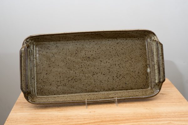 the photograph shows a clay coyote flameware fish tray resting on a stand. the stand allows the fish tray to be almost vertical, and allows the viewer to see the entire inside cooking surface of the tray. the tray and stand are sitting on a small light colored wooden table. the tray is glazed in coyote grey (grey with darker speckling through out the glaze). the background is a white wall. the photograph is lit with white light.