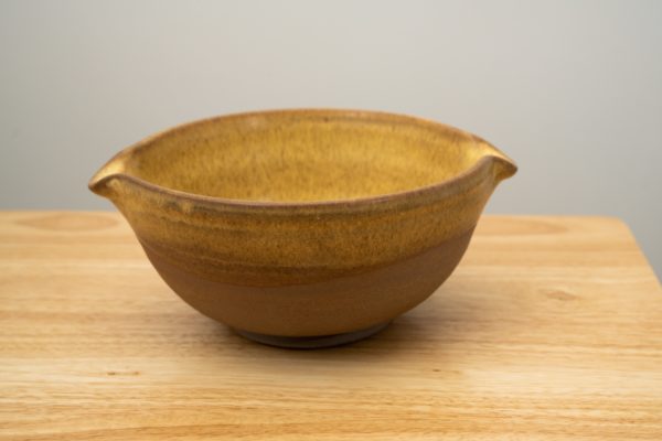 the photograph shows a clay coyote cassoulet bowl glazed in yellow salt resting on a small light colored wooden table. the cassoulet bowl has two spouts on opposite sides of the bowls lip. the yellow salt glaze covers the inside of the bowl completely and the upper portion of the outside. the outside bottom of the bowl is unglazed and shows off the natural color of the clay (reddish brown). the cassoulet bowl is resting flat on the small table. the background is a plain white wall. the photograph is lit with white light.