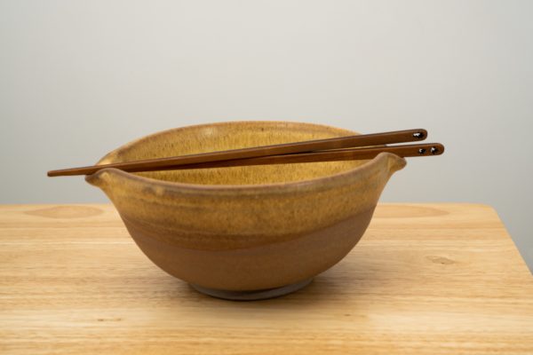 the photograph shows a clay coyote cassoulet bowl glazed in yellow salt resting on a small light colored wooden table. the cassoulet bowl has a pair of moonspoon brand chop sticks (sold at the clay coyote) resting on top. the chopsticks are resting in the pour spout grooves on top of the cassoulet bowl. the yellow salt glaze covers the inside of the bowl completely, and the upper part of the outside of the bowl. the lower outside portion of the bowl is unglazed, and shows off the natural color of the clay (reddish brown). the background is a plain white wall. the photograph is lit with white light.