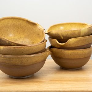 the photograph shows two stacks of three cassoulet bowls resting on a small light colored wooden table. the stack on the left side of the photograph is slightly closer to the camera then the stack on the right. both stacks contain 3 cassoulet bowls glazed in yellow salt. each bowl has the glaze cover the inside completely and the outside upper lip. the outside bottom of each bowl is unglazed and shows off the natural color of the clay (reddish brown). the background is a plain white wall. the photograph is lit with white light.