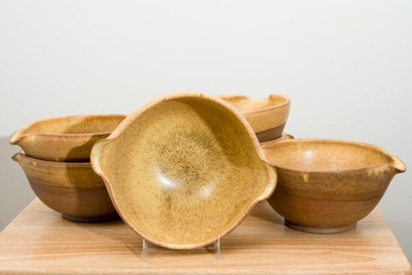 the photograph shows 6 clay coyote cassoulet bowls glazed in yellow salt resting on a small light colored wooden table. starting from the left of the photograph and working right the bowls are arranged: 2 cassoulet bowls stacked together, a cassoulet bowl on its side to show the inside of the bowls glaze patter, a stack of two cassoulet bowls then finally one cassoulet bowl by itself, resting flat on the table. the background is a plain white wall. the photograph is well lit with white light.