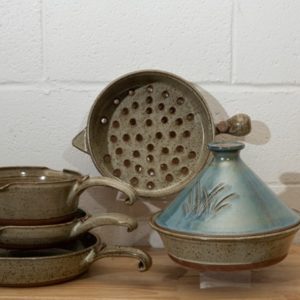 a horizontally framed photograph shows a clay coyote 5-piece set with a Mediterranean blue tagine. the 4 other flameware pieces that are not the mediterranean blue tagine are: clay coyote large skillet, clay coyote small skillet, clay coyote medium sauce pan and clay coyote grill basket. the grill basket is in the center of the photograph. the grill basket is on a clear plastic stand allowing it to stand on its side. it is on its side to allow full view of the holes in the bottom of the grill basket. the tagine in front and to the right of the grill basket. the tagine is on a small clear plastic stand so it is elevated off the wooden table. the tagine is at the grill baskets "5 o'clock" in terms of orientation. the three other flameware pieces are stacked to the left of the grill basket at the " 7 o'clock" position relative to the grill basket. the pans are stacked one on top of the other. the clay coyote large skillet is on the bottom, then the small skillet and finally the medium sauce pan. they are stacked with a small clear plastic riser under the small skillet and under the medium sauce pan, to allow a clearer view of each pan. the photograph is lit by overhead fluorescent lights