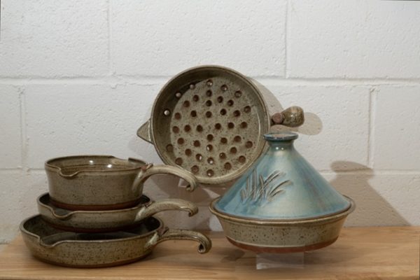 a horizontally framed photograph shows a clay coyote 5-piece set with a Mediterranean blue tagine. the 4 other flameware pieces that are not the mediterranean blue tagine are: clay coyote large skillet, clay coyote small skillet, clay coyote medium sauce pan and clay coyote grill basket. the grill basket is in the center of the photograph. the grill basket is on a clear plastic stand allowing it to stand on its side. it is on its side to allow full view of the holes in the bottom of the grill basket. the tagine in front and to the right of the grill basket. the tagine is on a small clear plastic stand so it is elevated off the wooden table. the tagine is at the grill baskets "5 o'clock" in terms of orientation. the three other flameware pieces are stacked to the left of the grill basket at the " 7 o'clock" position relative to the grill basket. the pans are stacked one on top of the other. the clay coyote large skillet is on the bottom, then the small skillet and finally the medium sauce pan. they are stacked with a small clear plastic riser under the small skillet and under the medium sauce pan, to allow a clearer view of each pan. the photograph is lit by overhead fluorescent lights