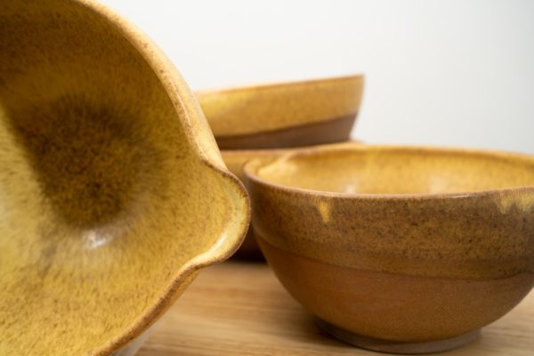 the photograph shows a close up view of 4 clay coyote cassoulet bowls glazed in yellow salt. in the back there are two bowls stacked one on top of the other. on the left side of the photograph a cassoulet bowl turned on it side to allow full view of the interior of the bowl. the cassoulet bowls are glazed fully on the inside of the bowl, and glazed on the outer upper lip of the bowl also. the outside bottom of the bowls are all unglazed. they show off the natural color of the clay (reddish brown). the background is a white wall. the photograph is lit with white light.