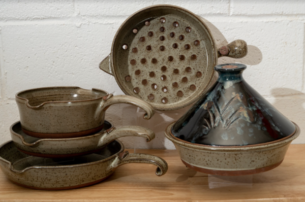 a horizontally framed photograph shows a clay coyote 5-piece set with a midnight garden tagine. the 4 other flameware pieces that are not the midnight garden tagine are: clay coyote large skillet, clay coyote small skillet, clay coyote medium sauce pan and clay coyote grill basket. the grill basket is in the center of the photograph. the grill basket is on a clear plastic stand allowing it to stand on its side. it is on its side to allow full view of the holes in the bottom of the grill basket. the tagine in front and to the right of the grill basket. the tagine is on a small clear plastic stand so it is elevated off the wooden table. the tagine is at the grill baskets "5 o'clock" in terms of orientation. the three other flameware pieces are stacked to the left of the grill basket at the " 7 o'clock" position relative to the grill basket. the pans are stacked one on top of the other. the clay coyote large skillet is on the bottom, then the small skillet and finally the medium sauce pan. they are stacked with a small clear plastic riser under the small skillet and under the medium sauce pan, to allow a clearer view of each pan. the photograph is lit by overhead fluorescent lights