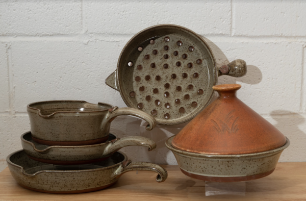 a horizontally framed photograph shows a clay coyote 5-piece set with a moroccan sand tagine. the 4 other flameware pieces that are not the moroccan sand tagine are: clay coyote large skillet, clay coyote small skillet, clay coyote medium sauce pan and clay coyote grill basket. the grill basket is in the center of the photograph. the grill basket is on a clear plastic stand allowing it to stand on its side. it is on its side to allow full view of the holes in the bottom of the grill basket. the tagine in front and to the right of the grill basket. the tagine is on a small clear plastic stand so it is elevated off the wooden table. the tagine is at the grill baskets "5 o'clock" in terms of orientation. the three other flameware pieces are stacked to the left of the grill basket at the " 7 o'clock" position relative to the grill basket. the pans are stacked one on top of the other. the clay coyote large skillet is on the bottom, then the small skillet and finally the medium sauce pan. they are stacked with a small clear plastic riser under the small skillet and under the medium sauce pan, to allow a clearer view of each pan. the photograph is lit by overhead fluorescent lights