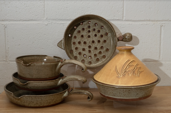 a horizontally framed photograph shows a clay coyote 5-piece set with a yellow salt tagine. the 4 other flameware pieces that are not the yellow salt tagine are: clay coyote large skillet, clay coyote small skillet, clay coyote medium sauce pan and clay coyote grill basket. the grill basket is in the center of the photograph. the grill basket is on a clear plastic stand allowing it to stand on its side. it is on its side to allow full view of the holes in the bottom of the grill basket. the tagine in front and to the right of the grill basket. the tagine is on a small clear plastic stand so it is elevated off the wooden table. the tagine is at the grill baskets "5 o'clock" in terms of orientation. the three other flameware pieces are stacked to the left of the grill basket at the " 7 o'clock" position relative to the grill basket. the pans are stacked one on top of the other. the clay coyote large skillet is on the bottom, then the small skillet and finally the medium sauce pan. they are stacked with a small clear plastic riser under the small skillet and under the medium sauce pan, to allow a clearer view of each pan. the photograph is lit by overhead fluorescent lights