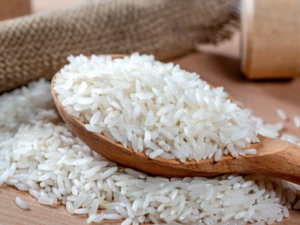 a horizontally framed photograph shows a close up view of the head of a wooden spoon filled with uncooked white rice. the spoon head is resting on more uncooked white rice. in the background out of focus is a rolled napkin or fabric placemat. everything in the photograph is white or shades of brown.