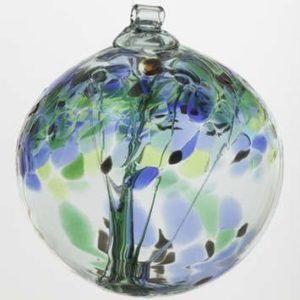 a vertically framed photograph shows a Kitras "encouragement" version of their hanging glass tree ornaments. The glass ball is photographed on an all white background with clear white light. there is nothing else in the photograph besides the glass ball. inside of the glass ball, the glass has been shaped to resemble a tree, and the upper half of the glass ball has been designed to look like a tree canopy. the encouragement style shown here dark blue, green and light green "leaves". at the top of the sphere there is small circular tube to hang the piece from.