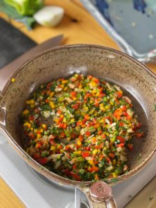 a vertically framed photograph shows a close up near overhead view of a clay coyote small skillet that is filled with diced vegetables. the skillet takes up almost the entirety of the photograph, yet the handle is still out of frame. the small skillet is sitting on a silver portable gas cooking burner. near the upper left corner of the photograph is part of a green pepper and part of a white onion. there is a part of a dark cutting cutting board and part of a large chefs knife visible. near the upper right corner is a clay coyote large tray, glazed in zappa. the tray is only partially visible with less then half of it in the photograph. the tray, the knife, the cutting board, the pepper and onion are all out of focus. the photograph is well lit with natural light