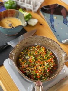 a vertically framed photograph shows a high angle, near overhead view of 3 pieces of clay coyote pottery being used to prepare an egg and diced vegetable dish. there is a clay coyote mixing bowl half full of beaten raw eggs resting on a black cutting board. the cutting knife is resting next to the mixing bowl and is also on the cutting board. the mixing bowl is near the upper left corner of the photograph. the bottom half of the photograph is taken up by the focal point of the photograph, a clay coyote small skillet filled with diced vegetables. the small skillet is resting on a silver portable cooking burner. near the upper right corner of the photograph a clay coyote large tray glazed in zappa can be partially seen. there isn't really anything in the tray, it appears to have a couple small pieces of diced vegetables in it. in between or near the mixing bowl and large tray are part of an onion, part of a green pepper, a whole green pepper and an out of focus egg carton. there are no eggs visible in the open egg carton.