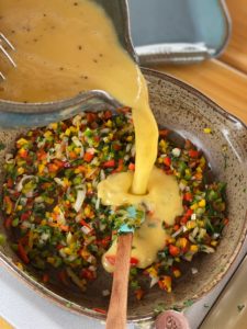 a vertically framed photograph shows an overhead view of a clay coyote mixing bowl pouring raw scrambled eggs into a clay coyote small skillet that is already partially filled with finely cut up vegetables. there are red, green and yellow peppers, onions, fresh herbs and tomatoes cut to very similar sizes. the egg mixture is in process of being poured into the vegetable mixture. there is a small amount of raw scrambled egg already in the small skillet, with a majority still in the mixing bowl. there is a wooden handled, teal silicone spatula sitting in the egg mixture that is forming in the small skillet. the silicone head of the spatula has some of the vegetable mixture the mixing bowl is only partially visible in the upper left corner of the photograph, with a lot of it cut out of frame. near the top center of the photograph is a clay coyote large tray. the tray is only partially visible, with only the lower right corner and handle visible.