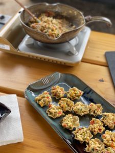 a vertically framed photograph shows a clay coyote large tray that is being used to serve cup shaped tortilla chips filled with an scrambled egg mixture. The tray is glazed in zappa, with only one handle visible, as one end is out of frame. the scrambled egg mixture has red and green vegetables mixed into the eggs, they are small enough that they are not easily identifiable. further away from the camera is a portable cooking burner. the burner has been used with a clay coyote small skillet to make the egg and vegetable mixture that is filling all the tortilla cups. there is a good amount of the scrambled egg mixture still in the small skillet. the large tray and the burner/small skillet are all sitting on a wooden picnic style table with large planks visible making up the table top. on the very left side of the photograph, mostly cut out of frame is a small stack of white paper napkins. the very corner of a clay coyote little dipper can be seen on top of the napkin pile. only one corner of the square style little dipper can be seen, as the rest is cut out of frame like the napkin pile.