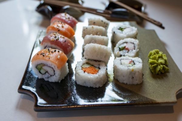 a horizontally framed photograph shows a high side angle view of a clay coyote sushi plate being used to serve sushi. there are 12 pieces of sushi on the sushi plate and two small dabs of wasabi. the sushi furthest to the left is nigiri style sushi with a piece of fish draped over each piece. there are tour like this. they alternate between (from top to bottom) tuna, salmon, tuna and salmon. each of these pieces of fish has had black and white sesame seeds sprinkled on them. the other pieces of sushi on the plate are regular style rolls. partially cut off on the top of the photograph is two clay coyote little dippers. each little dipper has a set of chopsticks resting across them. the little dippers are partially cut out of frame on the top edge of the photograph. they are also out of focus. the chop sticks are moon spoon chopsticks carried at clay coyote. the photograph is well lit with white natural light. the plate and the little dippers are all resting on a white counter top.