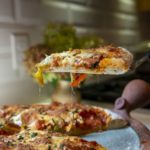 a vertically framed photograph shows clay coyote pizza stone that has bene used to make a fresh pizza. the pizza stone is only partially visible, as pizza is covering most of it. there is one slice of pizza being lifted just about 6 inches above the stone/rest of the pizza by a serving utensil. the hand holding the utensil is out of frame, and only a small amount of it is visible, as most is hidden under the pizza slice it is holding and the person holding it is completely out of frame. the pizza slice shows a good cross section of crust and toppings. there is a small amount of cornmeal visible on the crust, and on the pizza stone, beneath the lifted slice. in the background is a out of focus small clay coyote vase with a large bouquet of flowers resting in it.