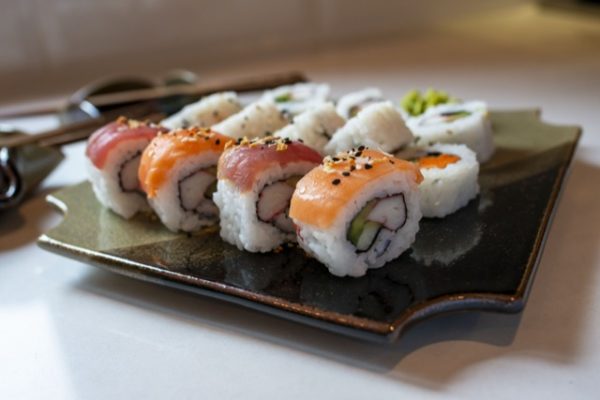 a horizontally framed photograph shows a low angle, almost side view of a clay coyote sushi plate being used to serve sushi. the sushi plate is glazed in mint chip. there are 12 pieces of sushi visible on the plate. the row of sushi closest to the viewer is 4 pieces of nigiri style sushi where the fish has been draped over the roll section. each piece of fish has had black and white sesame seeds sprinkled on them. the two rows of sushi behind this row are regular roll style, the "back" row of sushi and wasabi dabs next to it are out of focus. also out of focus and to the left of the sushi plate are two clay coyote little dippers. they both have chopsticks laying across them, but due to being out of focus and the camera angle, it is impossible to see if there is anything in them. most of the photograph is in shadow, but still well lit with natural white light. further down the white counter top a small section of yellow light can be seen, but it source is not photographed.