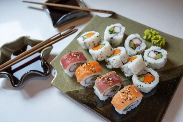 a horizontally framed photograph shows a high angle view of a clay coyote sushi plate and two little dippers being used to serve sushi. the partially cut out of frame on the top of the photograph is one of the little dippers. it has a set of moon spoon chopsticks resting across it from the top left corner to the bottom right. it has soy sauce poured in the bottom of the little dipper. below that little dipper and slightly to the right near the center left of the photograph is the other little dipper. this one is in the star or circular pattern and also has soy sauce poured in the bottom, with a set of moon spoon chopsticks resting across them. the clay coyote sushi plate is in the center of the photograph to the right center. the lower right corner of the square plate is cut out of frame. there is 13 pieces of sushi on the plate, along with two small dabs of wasabi. the sushi plate is glazed in mint chip. the bottom row of sushi is nigiri style where a piece of fish is draped over the roll. the fish alternates from left most to right with tuna, salmon, tuna, then another salmon. all 4 pieces of fish have had black and white sesame seeds sprinkled on them. the other pieces of sushi on the plate are regular roll style. the photograph is well lit with white light. both the little dippers and the sushi plate are all resting on a white counter top.