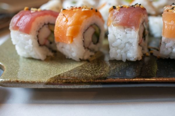a horizontally framed photograph shows a close up side view of a clay coyote sushi plate being used to serve sushi. the sushi plate is glazed in mint chip. the photographs angle is low, almost at the same height as the sushi. the sushi is nigiri style where the fish is draped on top of the roll pieces. the fish on top alternates from left to right with tuna, salmon, tuna then salmon again. each piece of fish has had black and white sesame seeds sprinkled onto them. there is more sushi behind this row, but it is out of focus and blocked by the nigiri style sushi in front of it. the clay coyote sushi plate is resting on a white counter top. the photograph is well lit with soft white light.