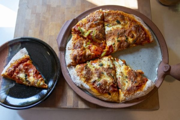 a horizontally framed photograph shows a overhead shot of a clay coyote pizza stone and a clay coyote sandwich plate, both with pizza on them. the pizza stone has been used to bake a fresh pizza, and one slice has been removed, and placed on the sandwich plate. the the clay coyote sandwich plate is glazed in mocha swirl. the pizza stones cooking surface is glazed in coyote grey to denote that it is flameware. the pizza stone and the sandwich plate are both resting on a square wooden butchers block. the butchers block is resting on a white kitchen counter top.