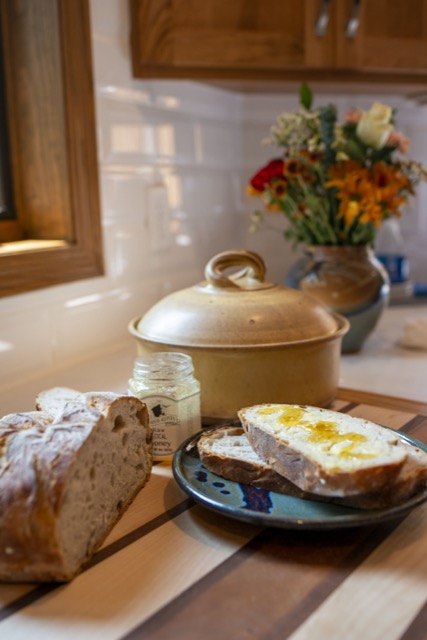 a vertically framed photograph shows a clay coyote bread baker in yellow salt with the lid on. in front of it, is a jar of honey with the lid off, a loaf of fresh bread that has had two slices cut off a placed on a clay coyote sandwich plate glazed in zappa. the two bread slices are stacked on top of each other. the top slice of bread has had honey drizzled and spread over it. all of this is sitting on a cutting board. behind the cutting board and further back into the background (and out of focus) is a large clay coyote vase glazed in joes blue with a large bouquet of flowers resting in it.